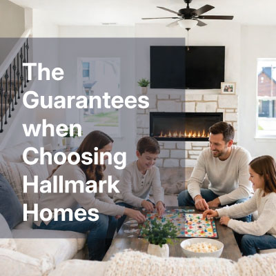 Family of four playing a board game at a cozy living room table with a stone fireplace and a mounted TV; overlay text reads 'The Guarantees when Choosing Hallmark Homes'