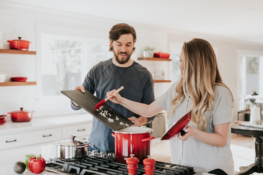 Couple cooking together in a bright kitchen; man holds a cutting board while woman adds chopped ingredients to a red pot on the stove, smiling at each other.